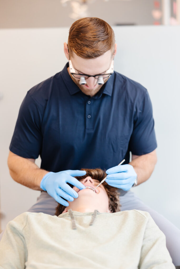 An orthodontist wearing magnifying loupes, a navy polo, and blue gloves examines a young male patient’s teeth using a dental scaler. The patient, dressed in a light green sweatshirt, is reclined in a black dental chair inside an orthodontic office.