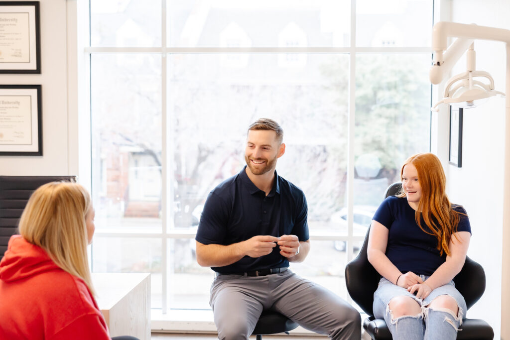 A smiling Dr. Bean in a navy polo shirt sits on a stool, talking with a young female patient and her parent in a bright dental office. The patient, a red-haired girl in a navy shirt and ripped jeans, sits in a dental chair, while her parent, wearing a red hoodie, listens attentively.