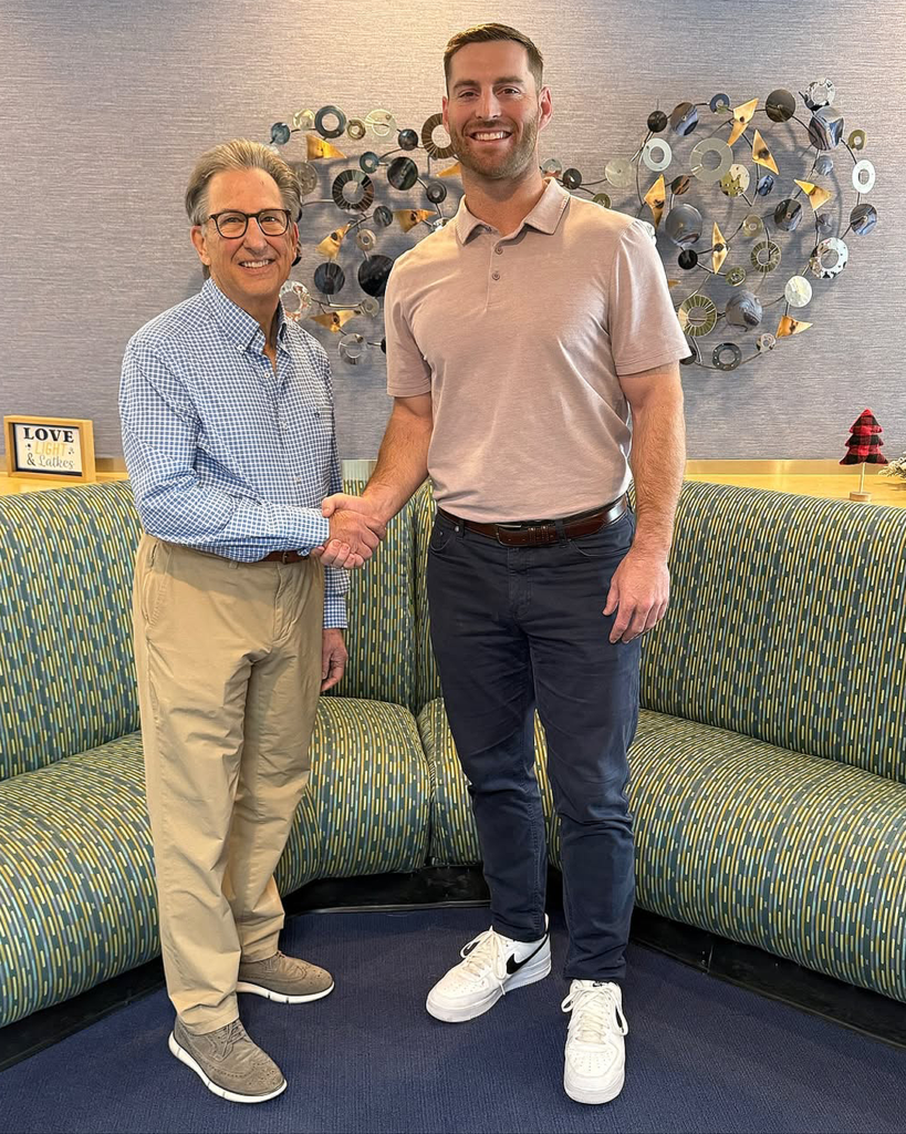 Two orthodontists, Dr. Geller on the left and Dr. Bean on the right, shaking hands and smile in a warmly decorated office. Dr. Geller is wearing glasses, a blue checkered shirt, and khaki pants, stands next to Dr. Bean, who is dressed in a light pink polo and dark pants. A decorative metal wall art piece and a cozy curved bench are visible in the background.