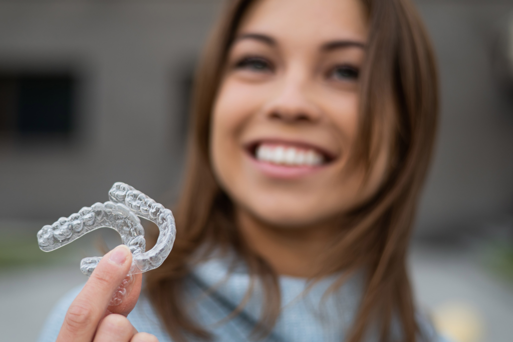 A young adult with medium-length brown hair smiles slightly out of focus in the background. In the foreground, their left hand holds up a clear, plastic upper and lower dental retainer or aligner, often associated with brands like Invisalign. The retainers are the main focus, held up against a blurred outdoor or neutral background. The image highlights this modern orthodontic appliance.