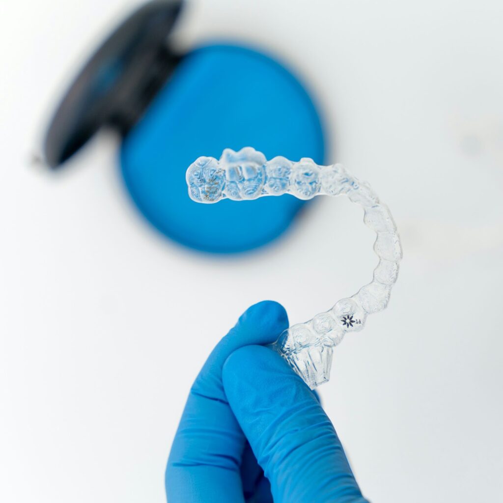 A gloved hand holding a clear, plastic retainer with a blue retainer case in the background.