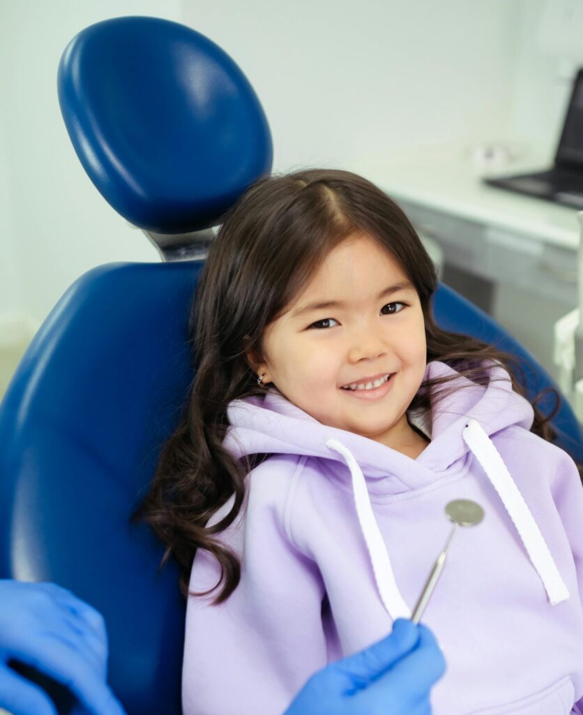 Young girl with long dark hair sitting in a dental chair, smiling confidently at the orthodontist holding a dental mirror. She’s wearing a lavender hoodie.