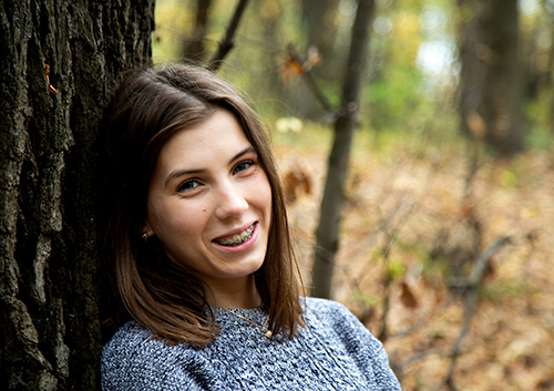 A young woman with brown hair and braces smiles while leaning against a tree in a forest setting.