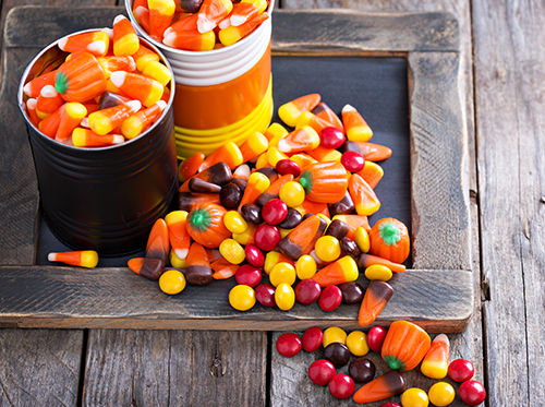 Assortment of Halloween candies, including candy corn, pumpkin candies, and colorful round candies, spilling from metal cans onto a wooden board.
