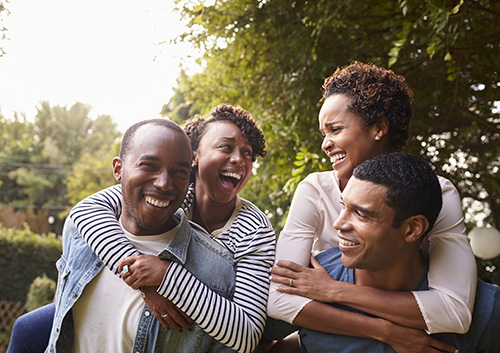 Four happy African American adults laughing and giving piggyback rides outdoors.