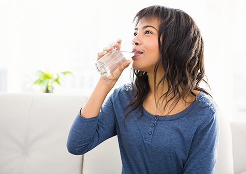 Young woman with dark hair drinking a glass of water while sitting on a couch.