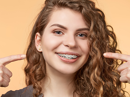 A smiling young woman with curly brown hair points to her clear braces, set against a light orange background.