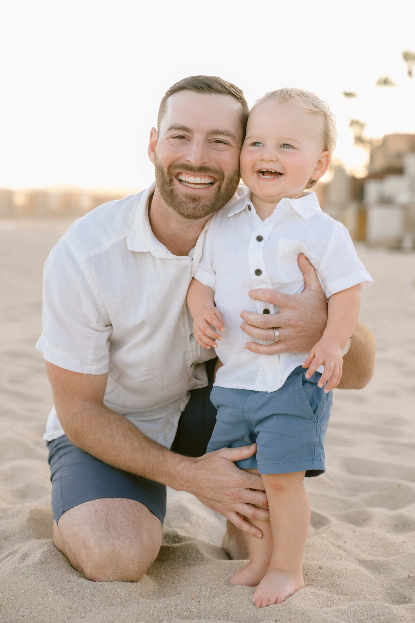 A father kneeling in the sand, smiling and hugging his laughing toddler son.