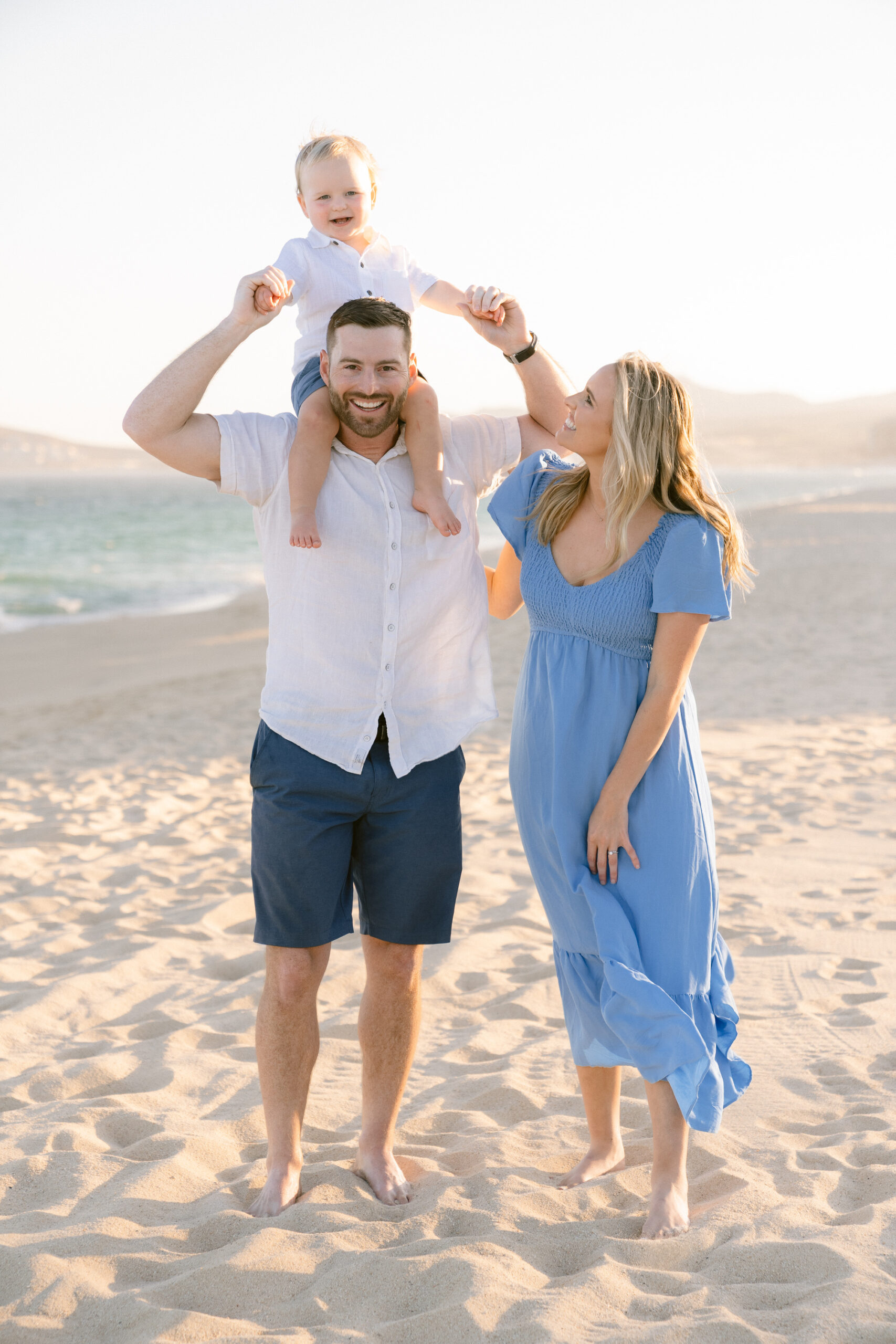 A father holding his toddler on his shoulders while the mother looks up at them, all smiling on the beach.