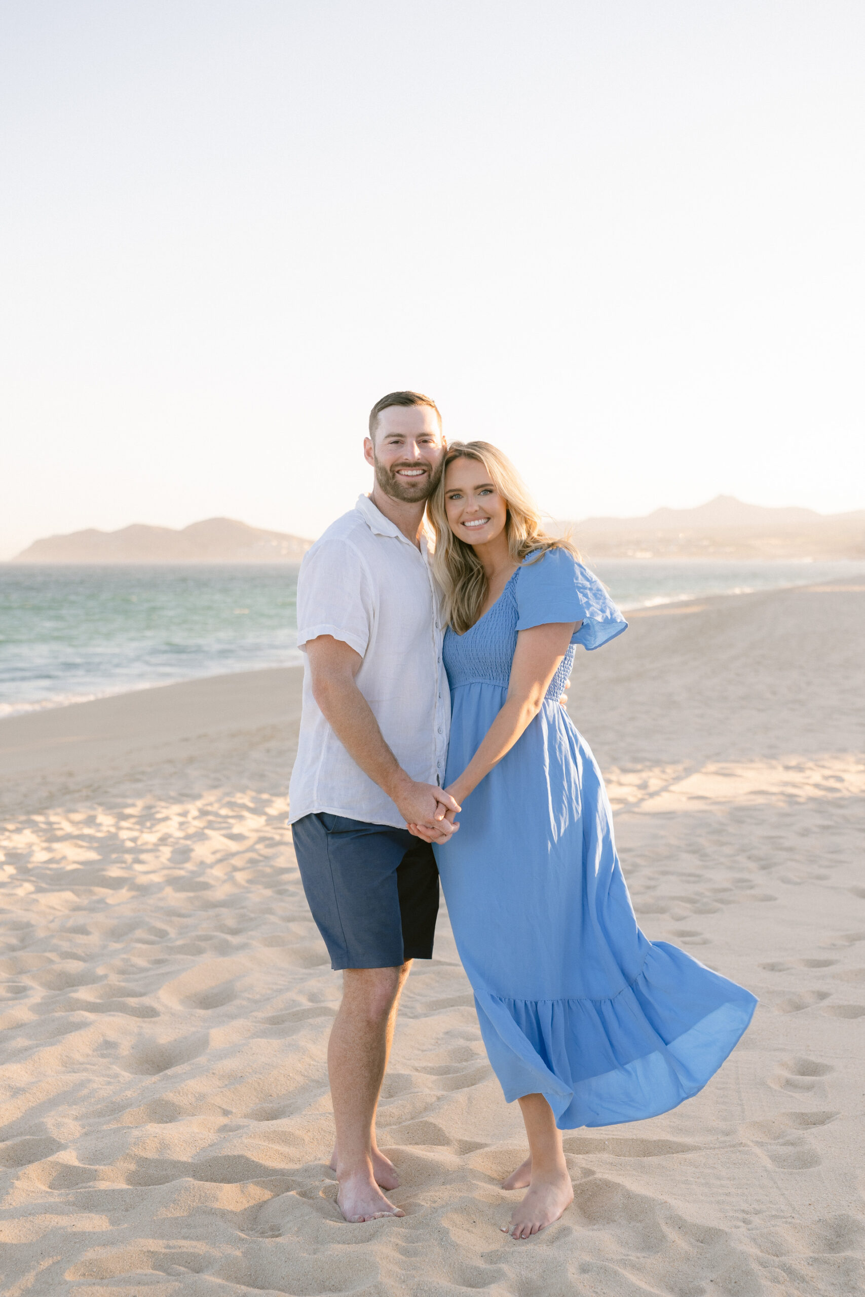 A smiling couple holding hands while standing on the beach at sunset.