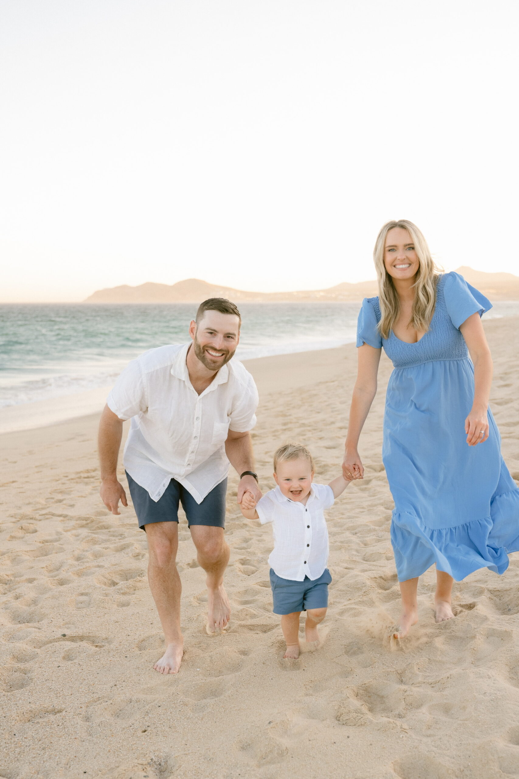 A smiling couple walking barefoot on the beach with their toddler between them, holding their hands.