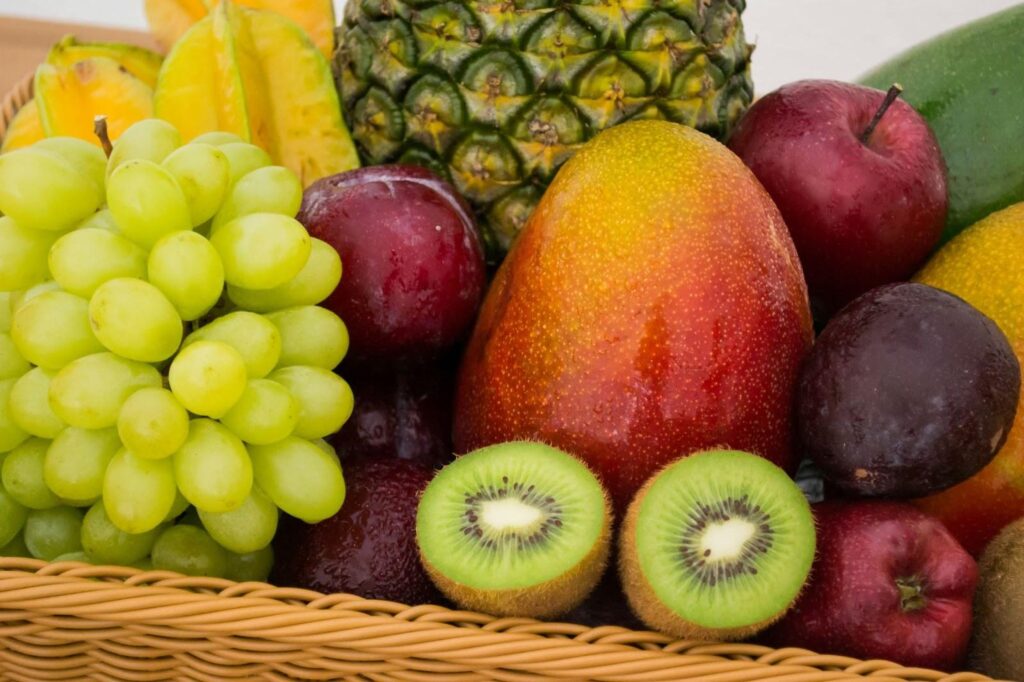 A woven basket filled with colorful fresh fruits, including grapes, kiwis, mango, apples, pineapple, and starfruit.