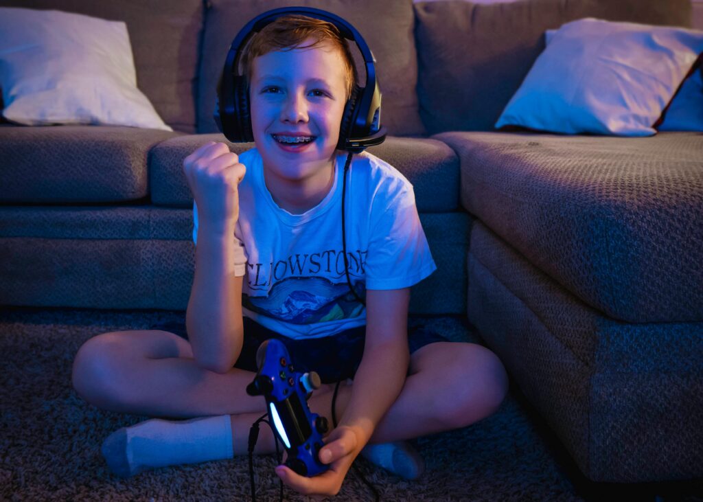 A smiling boy wearing braces and a headset sits on the floor playing a video game.