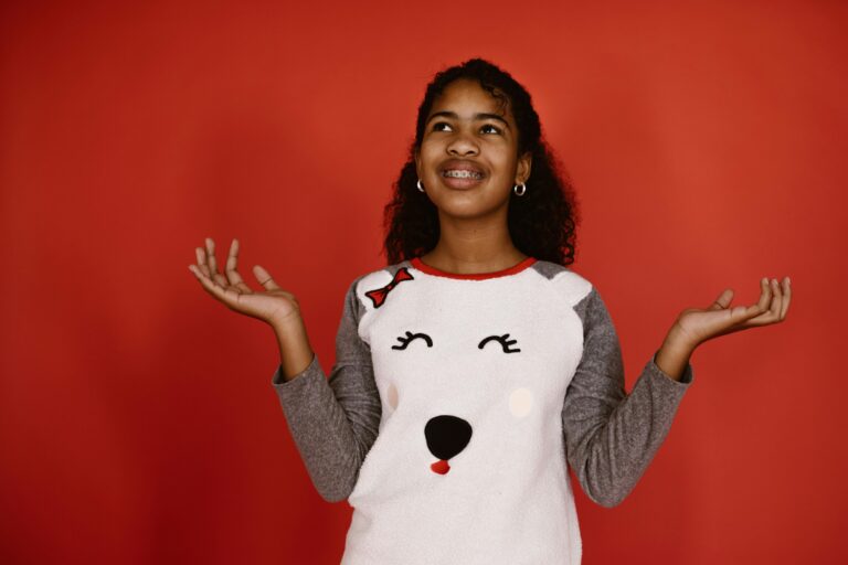 A girl with braces in a bear sweatshirt stands against a red background, arms out, looking up.
