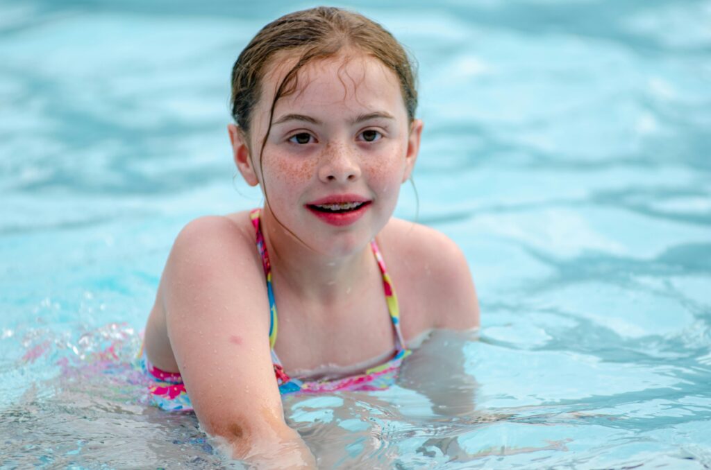 A girl with braces and wet hair smiles at the camera while in a swimming pool.