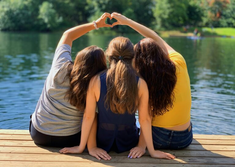 Three young women sit side-by-side on a wooden dock overlooking a calm lake, with two forming a heart shape above their heads on a summer day.
