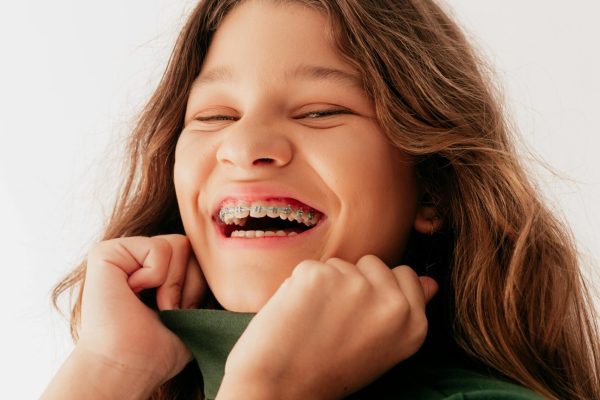 Smiling young girl with long hair wearing braces and a green shirt, pulling her collar playfully while laughing against a plain white background.