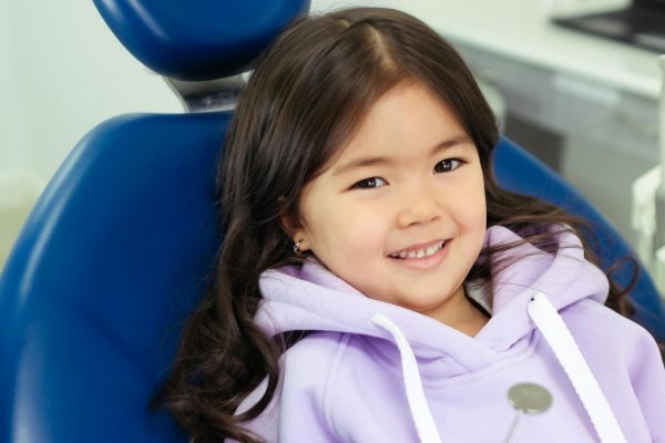Young girl with long dark hair sitting in a dental chair, smiling confidently at the orthodontist holding a dental mirror. She’s wearing a lavender hoodie.