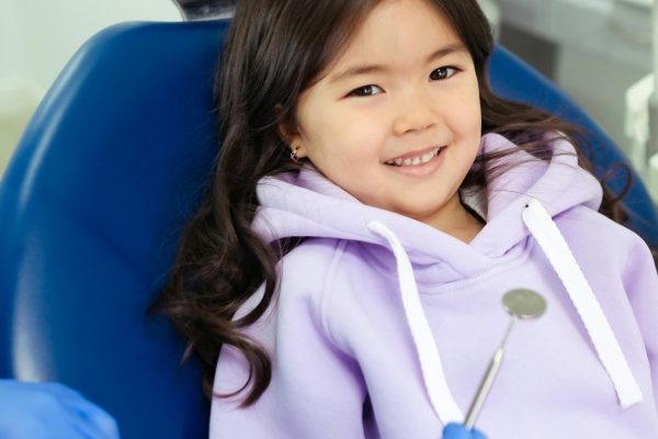 Young girl with long dark hair sitting in a dental chair, smiling confidently at the orthodontist holding a dental mirror. She’s wearing a lavender hoodie.